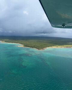 Rain falling over a forested coastline, seen from under the wing above turquoise water.